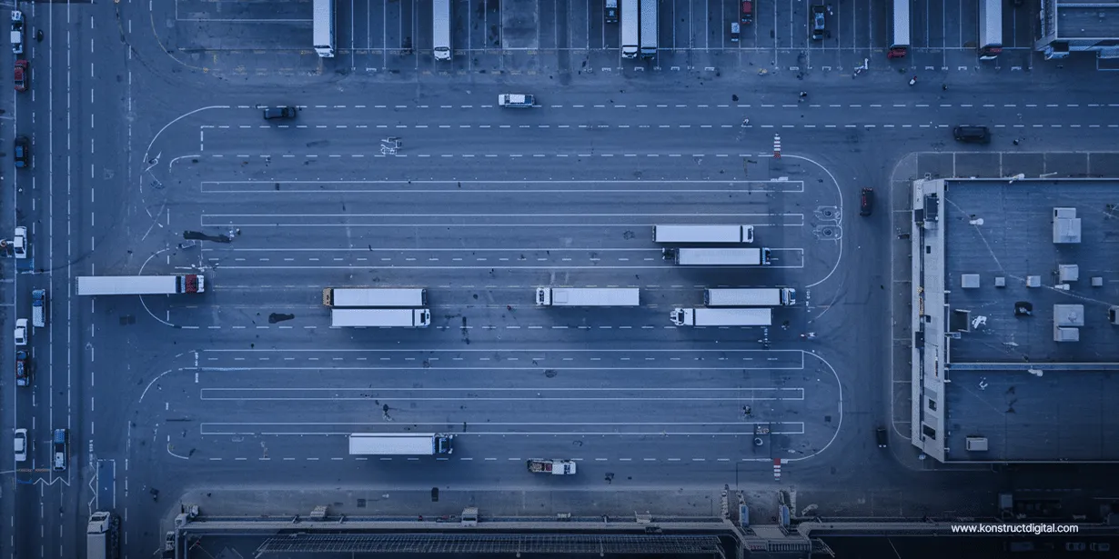 Aerial view of a logistics hub with multiple semi-trucks maneuvering and parked across wide lanes and loading bays, surrounded by warehouses and industrial buildings