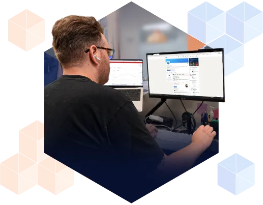 A person sits at a desk with two monitors, focused on a computer screen displaying a social media page. The room has a tech-themed decor.