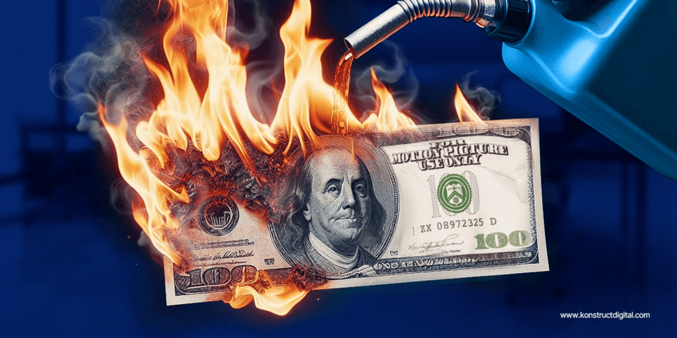 A US 100-dollar bill being consumed by flames. The fire appears to be fed by a jerry can pouring gasoline onto the blaze. The background is a blurred blue hue that contrasts with the bright orange and yellow of the fire, drawing the eye to the incineration of the currency. There's a watermark that reads "www.konstructdigital.com" at the bottom right. The image could be illustrating the concept of "burning money" or financial waste.