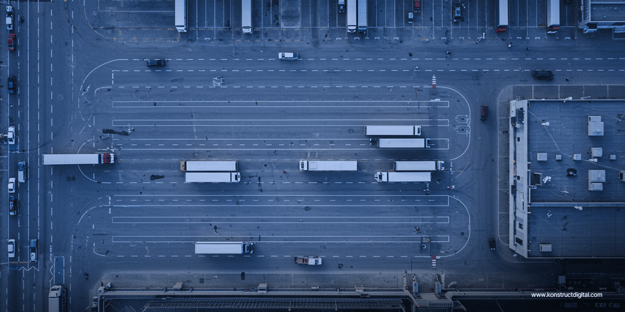 Aerial view of a logistics hub with multiple semi-trucks maneuvering and parked across wide lanes and loading bays, surrounded by warehouses and industrial buildings